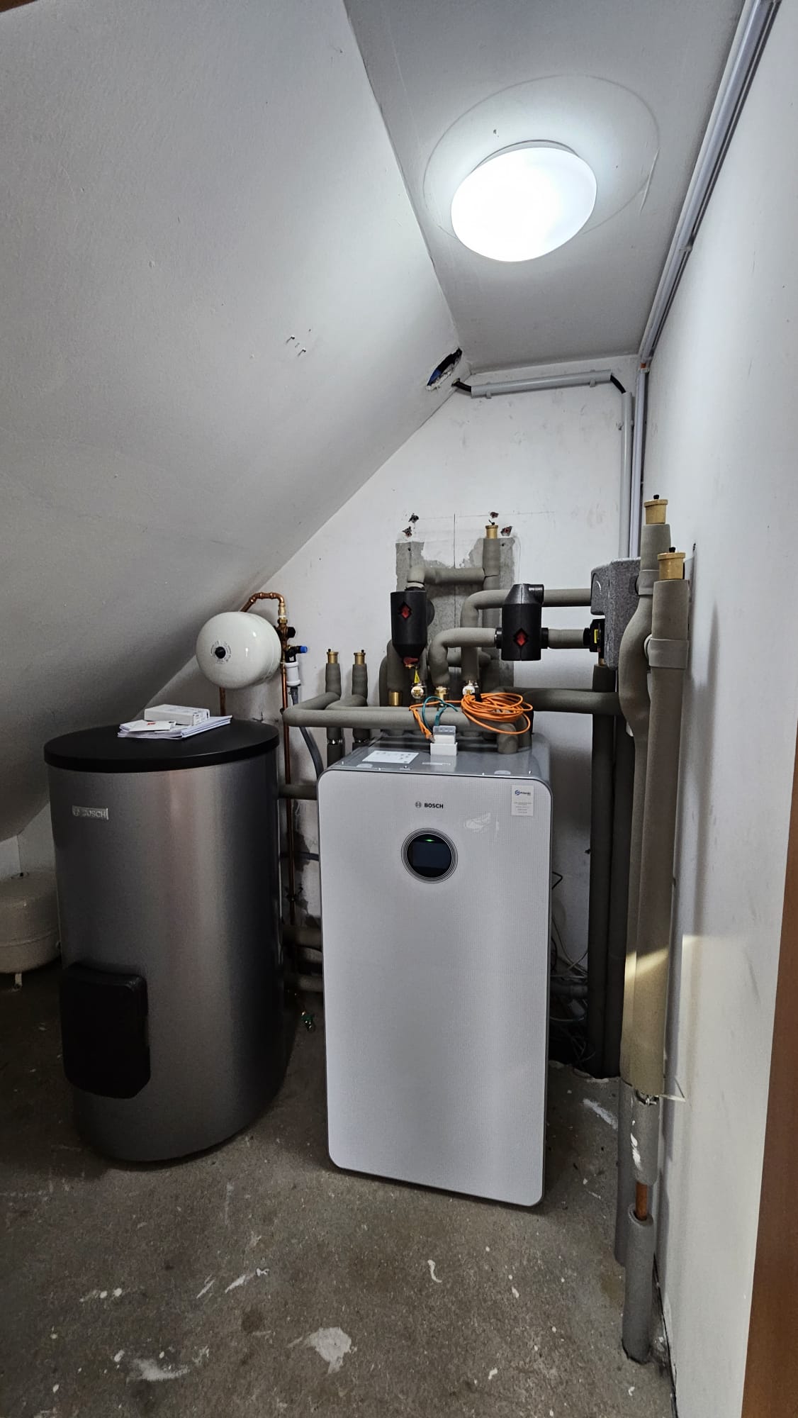 Eine Bosch-Wärmepumpen-Inneneinheit steht kompakt im Heizungskeller, angeschlossen an Speicher und umfangreiche Rohrleitungen.